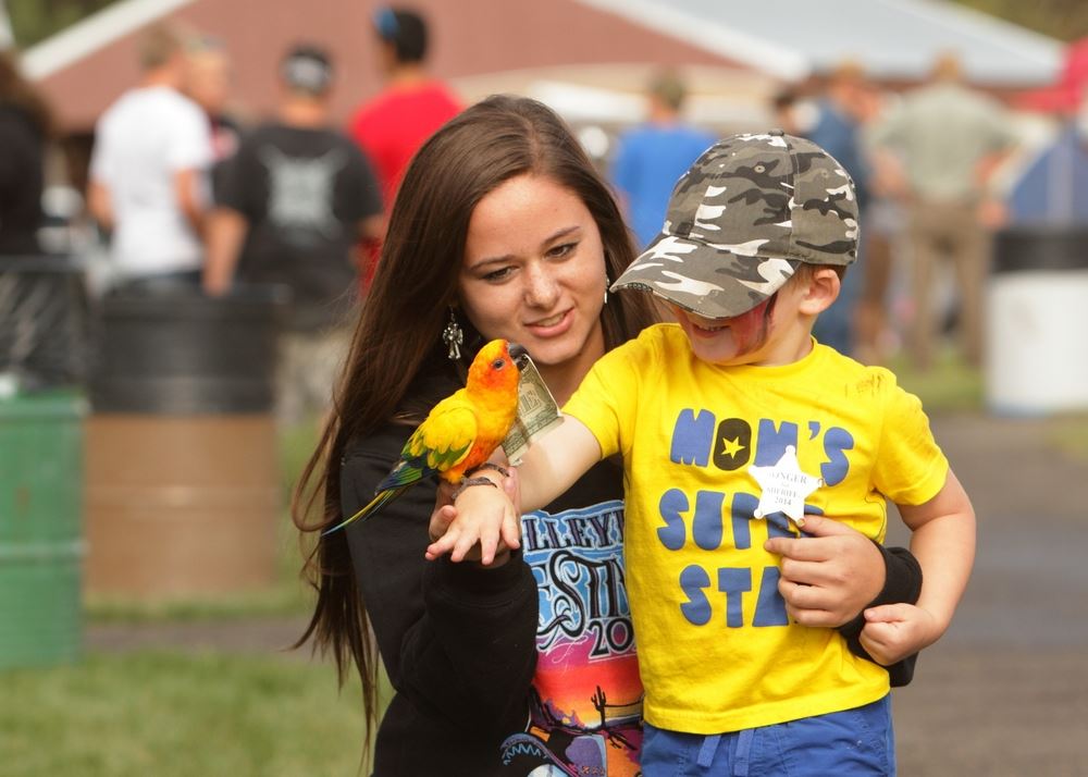 A little boy smiles at a parrot sitting on his arm holding a dollar bill in its beak.
