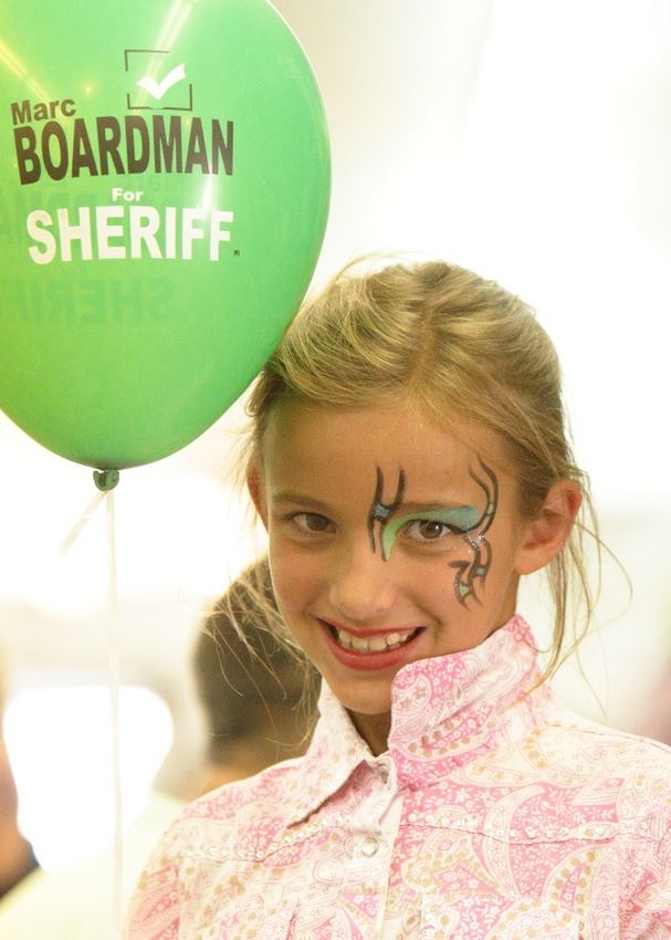 A little girl with her face painted holds a green balloon.