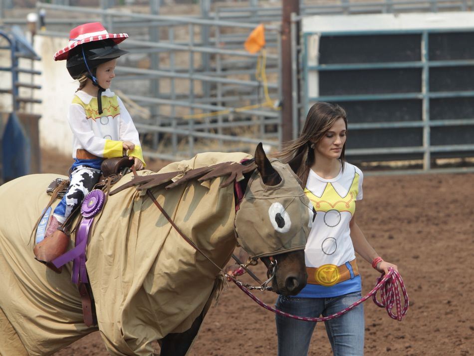 Woman walks a horse dressed as Bullseye, carrying a little girl dressed as Jessie from Toy Story 2.